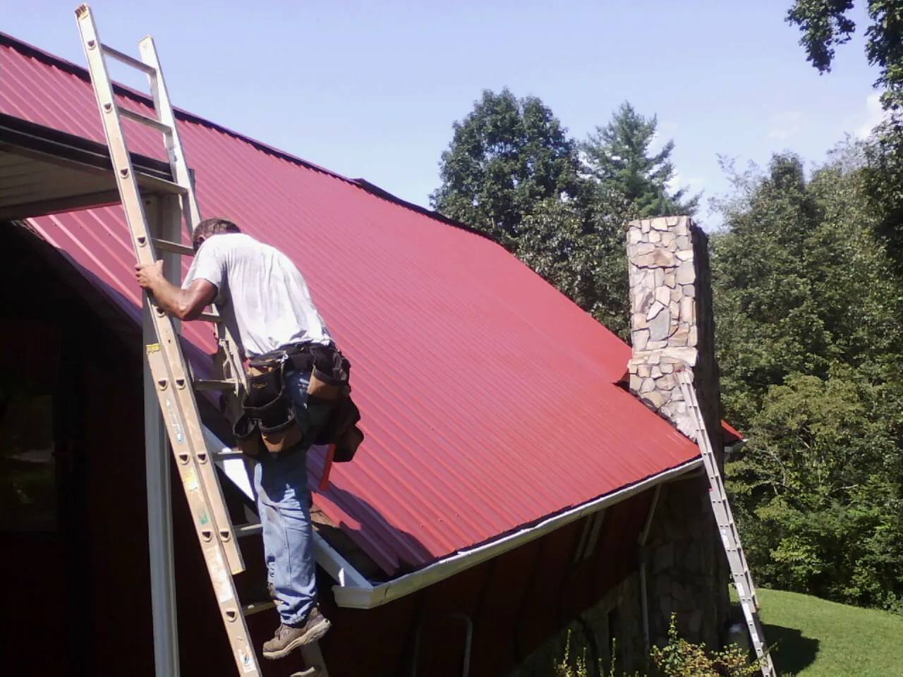 Roofer on a ladder beside a red metal roof and stone chimney — Soffit & Fascia Repair by Roofing Brothers in Wahoo